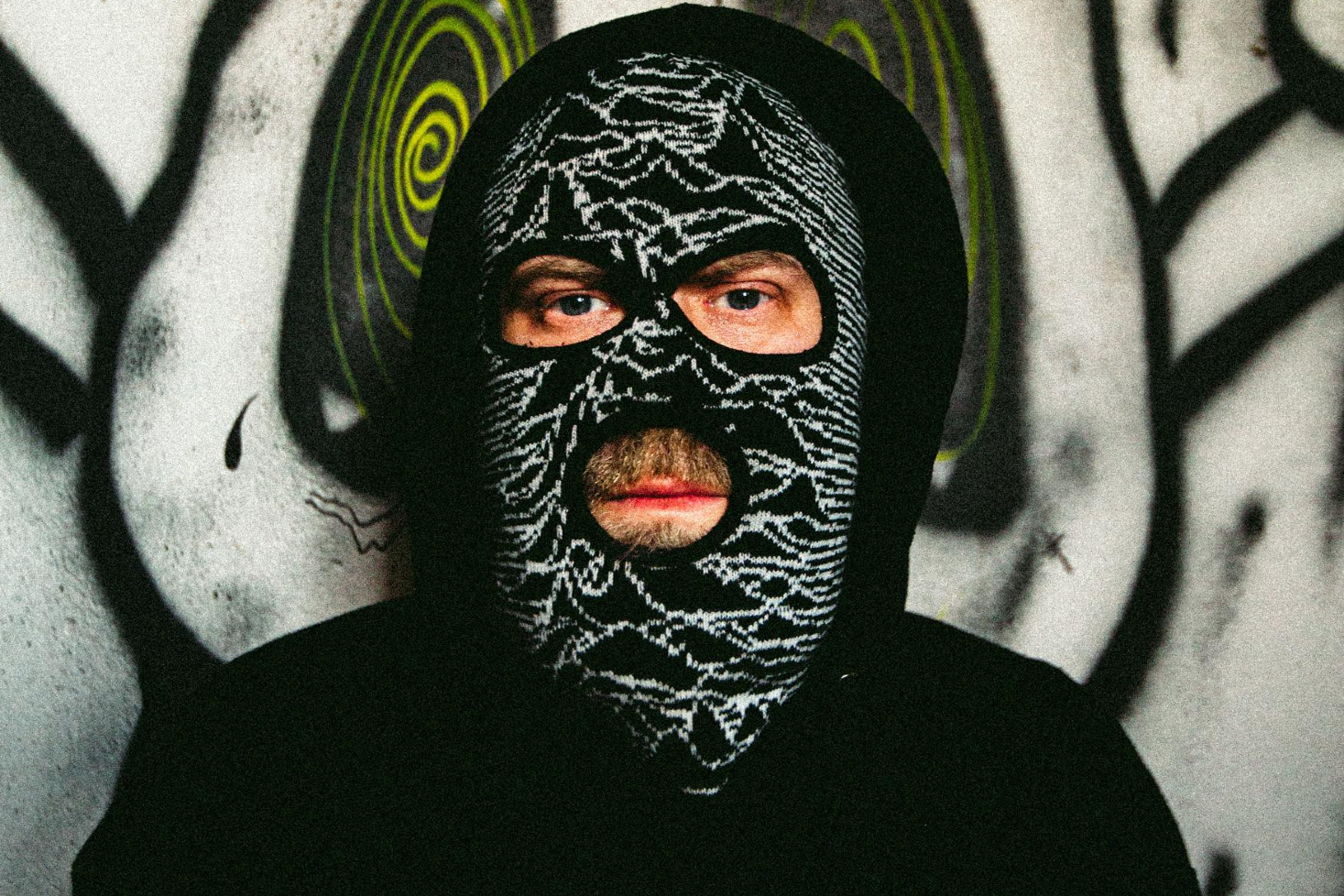 Eevil Stöö esiintyy mustassa suojamaskissa abstraktin graffitiseinän edessä, korostaen hänen mystistä ja omaleimaista tyyliään.