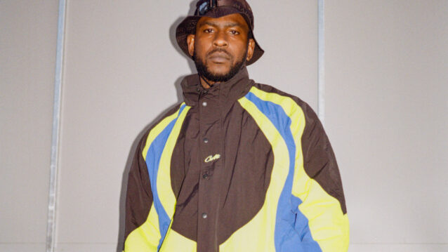 Skepta seisoo katsoen kameraan tuulitakki päällä ja lierihattu päässä.