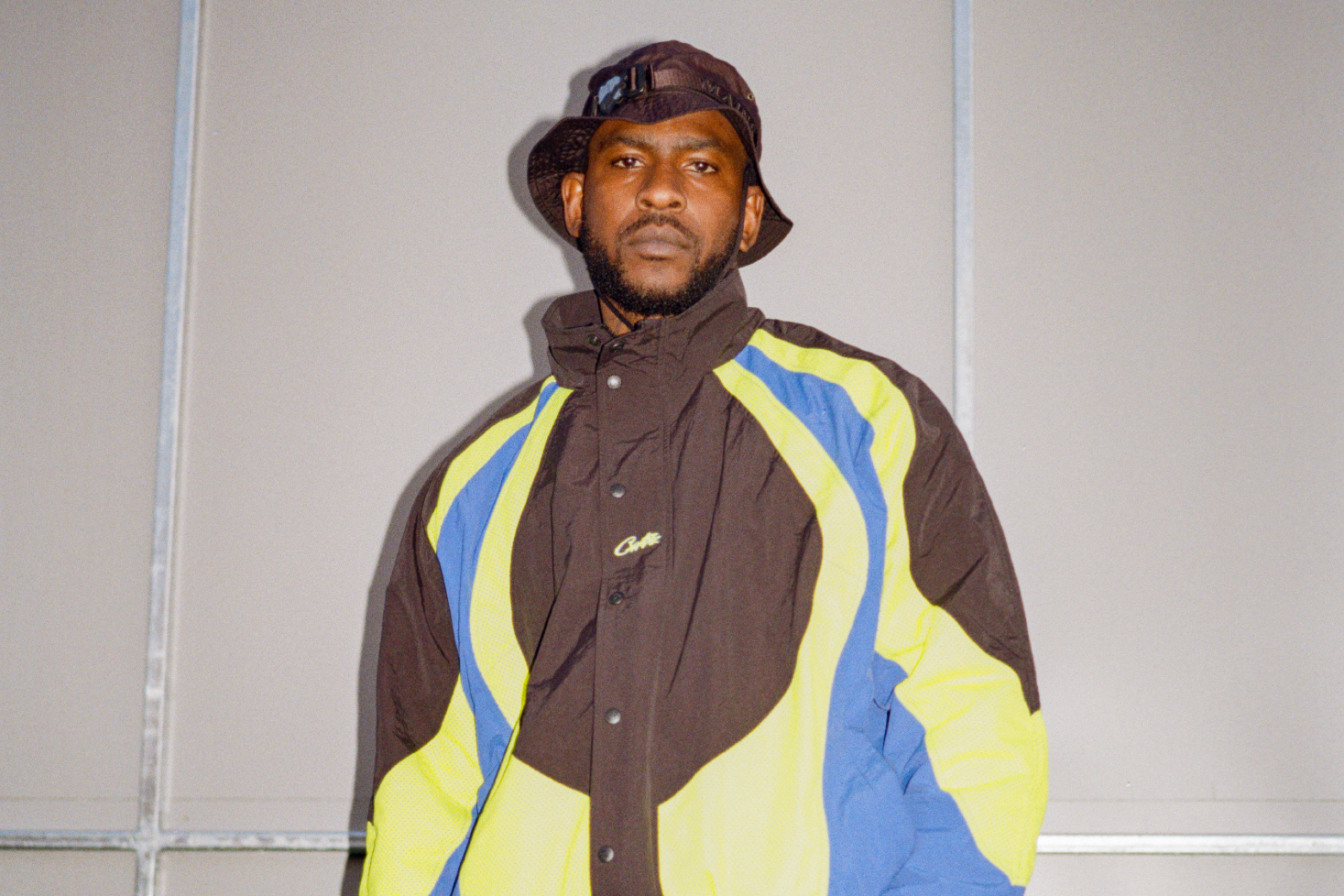 Skepta seisoo katsoen kameraan tuulitakki päällä ja lierihattu päässä.