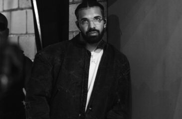 Räppäri Drake mustavalkoisessa kuvassa. Drakella on päällään tumma takki ja silmälasit, ja nojaa kuvassa seinään.