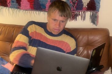 Räppäri AB kuvattuna sohvalla kahden tietokoneen kanssa, päällään raidallinen villapaita. Klangin haastattelu pyrkii selvittämään vastauksen kysymykseen: Kuka on AB?