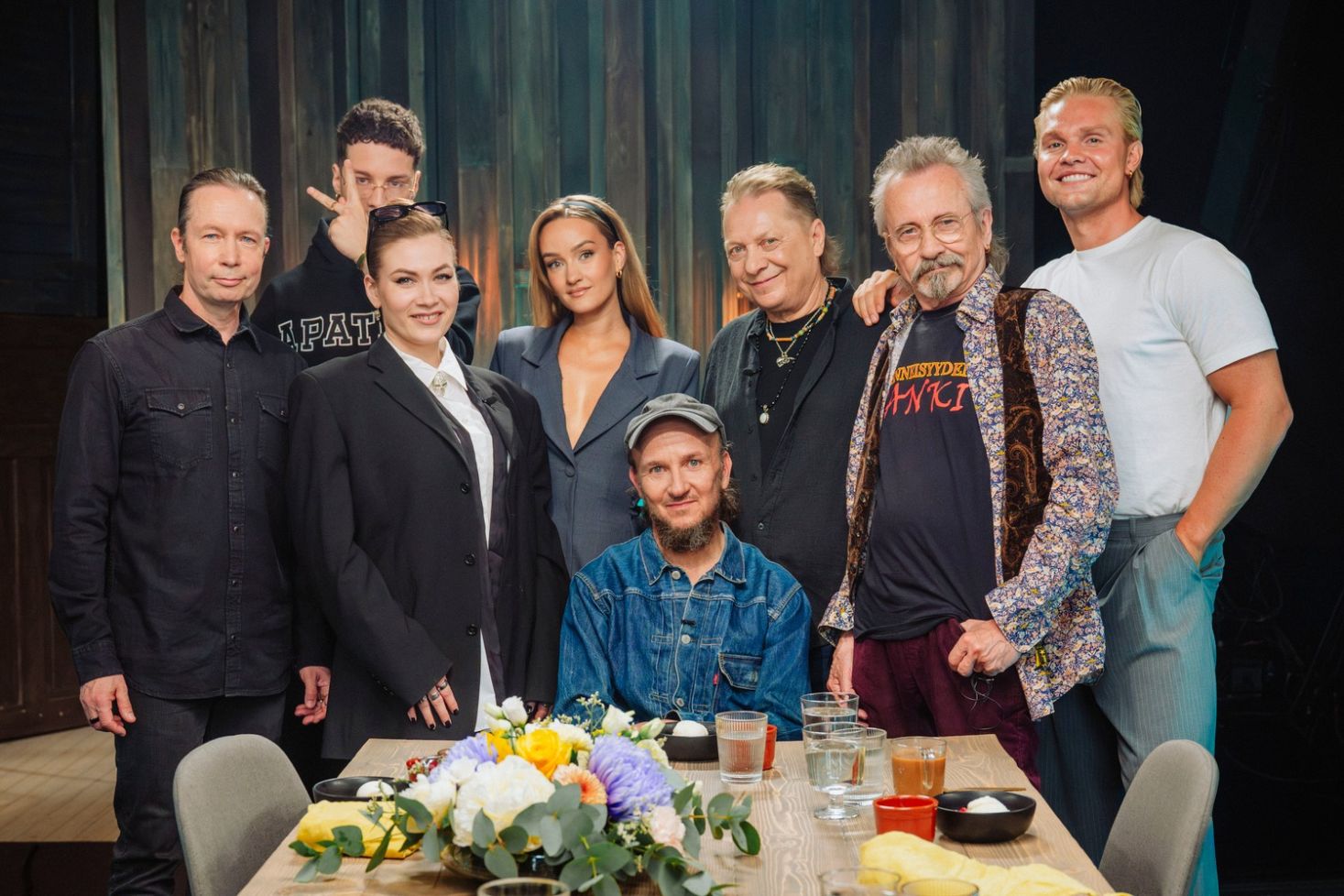 Samuli Putron Vain elämää -jakson artistit poseeraavat kukkakoristellun pöydän ääressä studiossa.